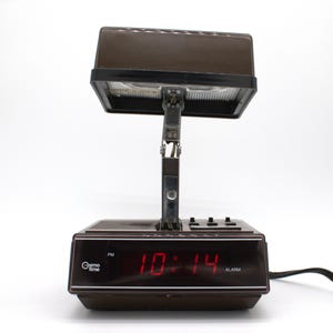May include: A brown Cosmo Time digital alarm clock with a rectangular base and a hinged, adjustable lamp head. The front display features a red digital clock showing 10:14 PM, with the brand name Cosmo Time and the word ALARM printed in white text.