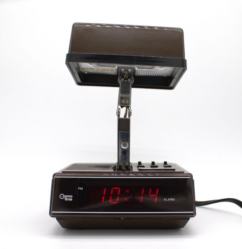 May include: A brown Cosmo Time digital alarm clock with a rectangular base and a hinged, adjustable lamp head. The front display features a red digital clock showing 10:14 PM, with the brand name Cosmo Time and the word ALARM printed in white text.