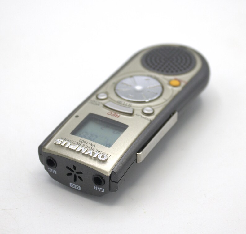 May include: A silver Olympus VN-1800 digital voice recorder with a black clip. The recorder has a small LCD screen, a record button, and a speaker.
