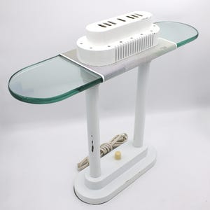 May include: A white desk lamp with a unique design. It features a rectangular base, two vertical supports, and a horizontal top with two clear glass shelves. The lamp has a white finish and a power cord with a switch.