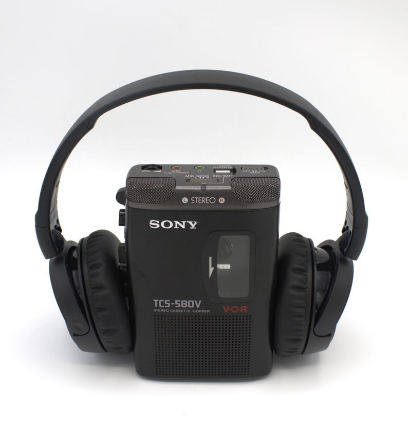 May include: A black Sony TCS-580V stereo cassette-corder with attached black headphones. The cassette-corder has a speaker, microphone, and various control buttons. The headphones arch over the top of the cassette-corder.
