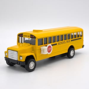 May include: A bright yellow school bus toy with black tires and a white stop sign. The words "SCHOOL BUS" are printed in black on the side. The bus has a detailed front grill and windows.
