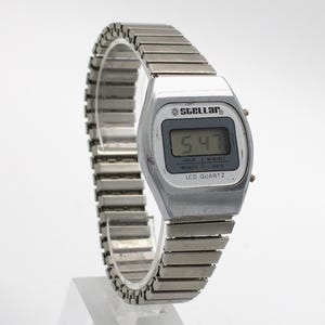 May include: A silver-toned digital wristwatch with a metallic expansion band. The watch face features the brand name Stellar, a digital display showing 5:47, and the text LCD QUARTZ, HOUR, MIN/SEC, MONTH, and DATE printed on the screen border.