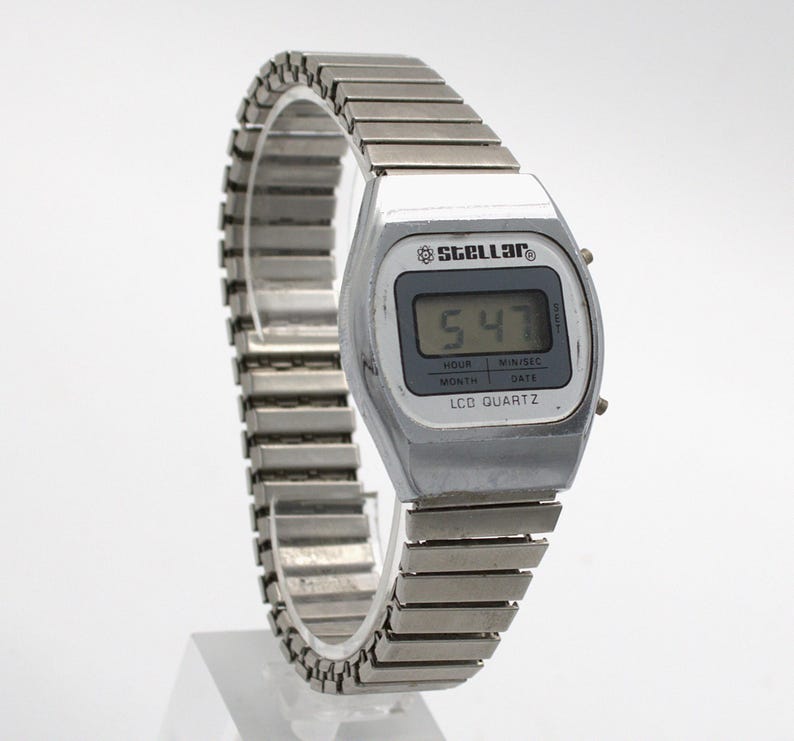 May include: A silver-toned digital wristwatch with a metallic expansion band. The watch face features the brand name Stellar, a digital display showing 5:47, and the text LCD QUARTZ, HOUR, MIN/SEC, MONTH, and DATE printed on the screen border.