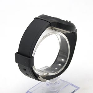 May include: A black digital wristwatch with a rectangular face and a black rubber strap. The watch is displayed on a clear plastic stand, showcasing the band's design and clasp. The watch face has small buttons.