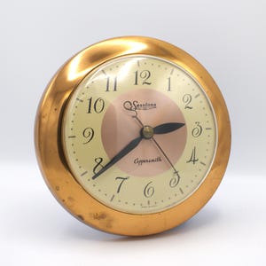 May include: A vintage Sessions Coppersmith clock with a gold-tone metal casing. The clock face is cream-colored with black numerals and hands. The center features a pink circle with the Sessions logo. The clock is a classic, retro design.