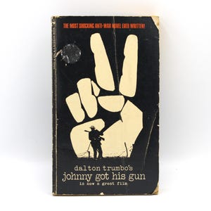 May include: Vintage paperback book cover for "Johnny Got His Gun" by Dalton Trumbo. The cover features a black background with a large, off-white peace sign. The title and author's name are also displayed in off-white, along with the text "is now a great film."