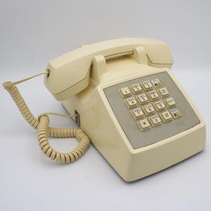 May include: Cream-colored AT&T push-button telephone with a handset and coiled cord. The phone has a rectangular keypad with numbered buttons and a gray panel. The phone is a vintage design.