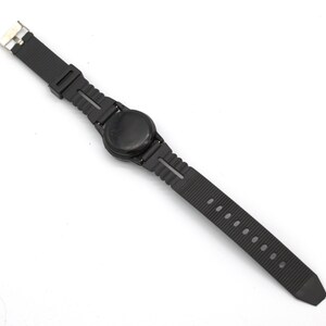 May include: A black wristwatch with a round face and a textured rubber strap. The watch has a simple, minimalist design with a metal clasp. The strap has multiple holes for adjustment.