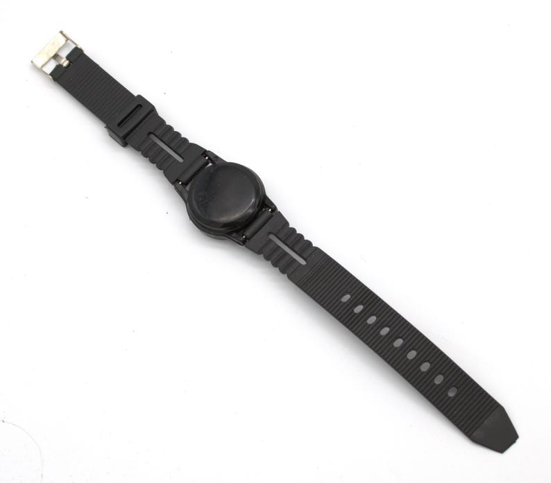 May include: A black wristwatch with a round face and a textured rubber strap. The watch has a simple, minimalist design with a metal clasp. The strap has multiple holes for adjustment.
