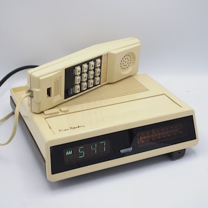 May include: A vintage Pierre Cardin telephone and radio alarm clock combo in cream and black. The phone has a handset with a keypad, and the clock displays the time as 5:47 AM. The radio dial is visible below the clock display.