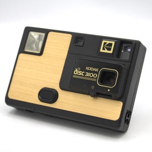 May include: A vintage Kodak Disc 3100 camera with a black body and a light wood-grain finish on the front. The camera features a flash, a lens, and the Kodak logo. The text "KODAK disc 3100" is printed on the front.