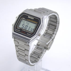 May include: A silver-toned digital watch with a rectangular black face displaying the time. The watch has a metal band and features the brand name "SKMEI". The watch face also includes the text "ALARM CHRONOGRAPH", "WATER 30M", and "RESIST".