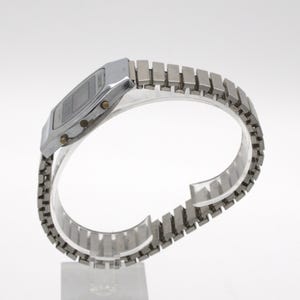 May include: A silver-toned digital wristwatch featuring a rectangular face with a dark display screen and a metallic expansion band. The watch case includes three small circular buttons on the side for adjusting settings and time functions.