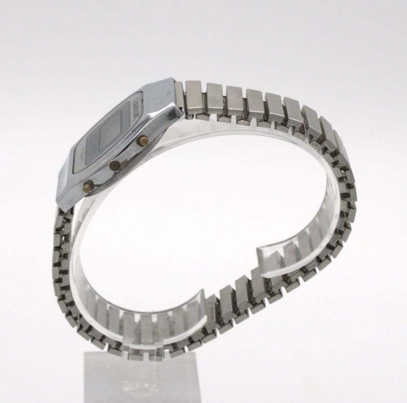May include: A silver-toned digital wristwatch featuring a rectangular face with a dark display screen and a metallic expansion band. The watch case includes three small circular buttons on the side for adjusting settings and time functions.