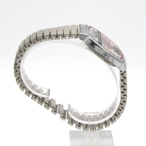Peut inclure: Une montre num&eacute;rique vintage avec un bracelet et un bo&icirc;tier en m&eacute;tal argent&eacute;. Le cadran de la montre a un affichage rouge et noir. Le bracelet est compos&eacute; de petits maillons rectangulaires. La montre est pr&eacute;sent&eacute;e sur un support incurv&eacute; transparent.