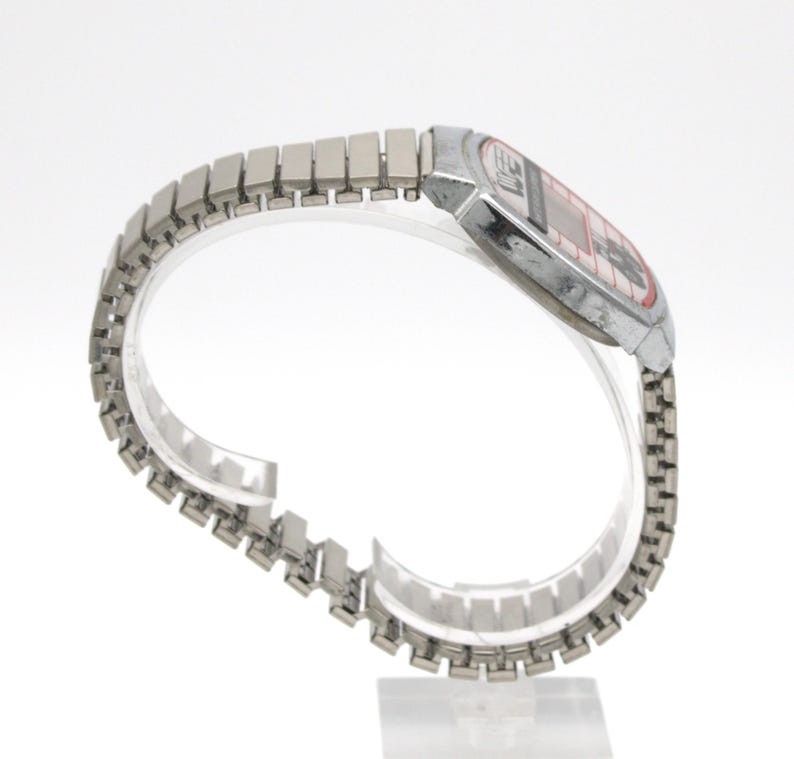 Peut inclure: Une montre num&eacute;rique vintage avec un bracelet et un bo&icirc;tier en m&eacute;tal argent&eacute;. Le cadran de la montre a un affichage rouge et noir. Le bracelet est compos&eacute; de petits maillons rectangulaires. La montre est pr&eacute;sent&eacute;e sur un support incurv&eacute; transparent.