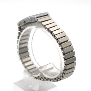 May include: A silver-toned digital wristwatch featuring a rectangular case and a flexible, expandable metal link bracelet. The side of the watch case displays two small circular buttons, and the metallic finish has a smooth, brushed texture.