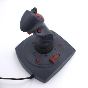 Puede incluir: Un joystick gris oscuro con botones y detalles en rojo. El joystick tiene una parte superior redondeada y una base con botones adicionales y una palanca de control. Un cable negro se extiende desde la base.
