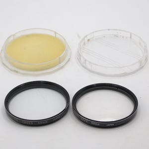 May include: Two black 55mm camera lens filters with clear glass, alongside two clear plastic cases. One case contains a yellow foam insert. The filters have text that reads "CANON CROSS 55mm JAPAN" and "UV+2 55mm JAPAN".