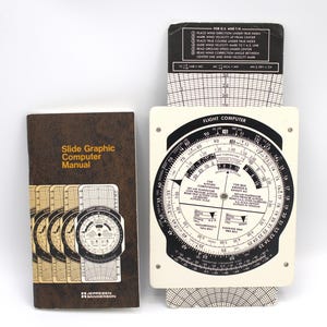 May include: A vintage flight computer set, including a slide graphic computer manual and a white and black flight computer. The manual is brown with the title "Slide Graphic Computer Manual". The flight computer has a circular dial with various scales.