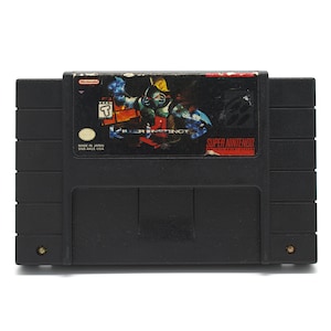 May include: Black Super Nintendo game cartridge for "Killer Instinct." The label features a colorful illustration of a character and the game title. The cartridge is marked "Made in Japan" and "SAS-ALE USA."