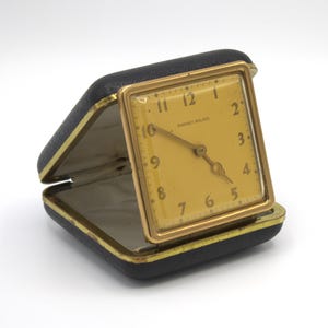 May include: A vintage travel clock with a gold-colored square face and hands, set in a black leatherette case. The clock face has the text "Punnet-Walker". The case is open, revealing a cream-colored interior.