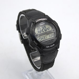 May include: A black Casio digital watch with a black fabric band. The watch face displays the time and other functions. The watch has the word "ILLUMINATOR" above the digital display.