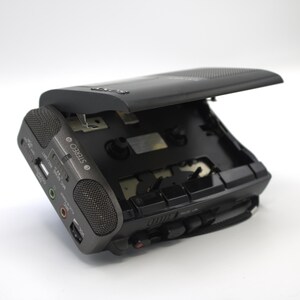 May include: A black portable cassette player with the top open, revealing the tape compartment. The device has a built-in speaker, USB port, and various input/output jacks. The word "STEREO" is printed on the side. A black carrying strap is attached.