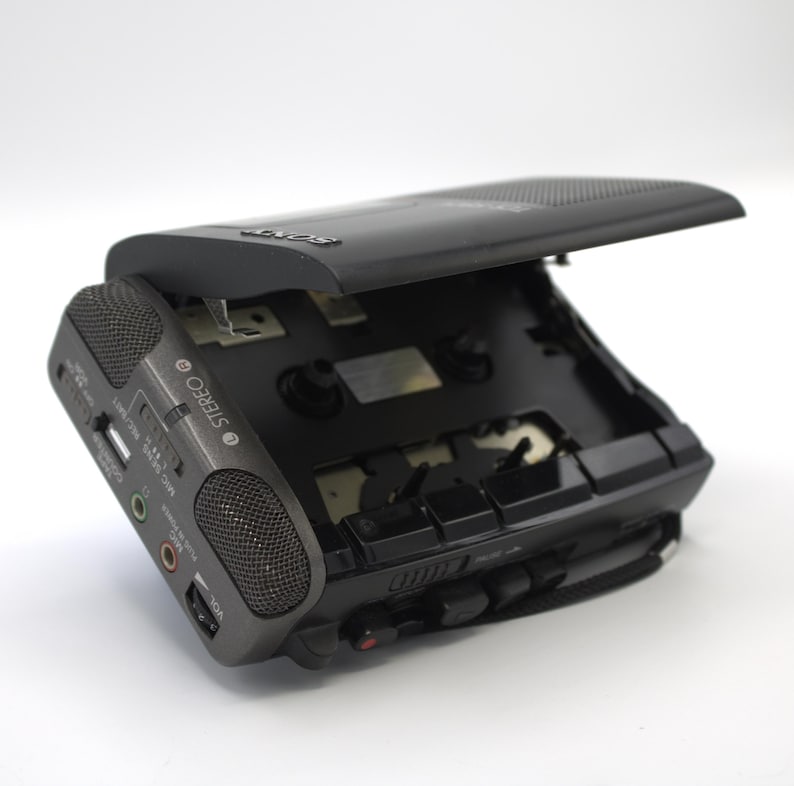 May include: A black portable cassette player with the top open, revealing the tape compartment. The device has a built-in speaker, USB port, and various input/output jacks. The word "STEREO" is printed on the side. A black carrying strap is attached.