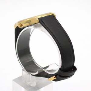 May include: A gold-toned wristwatch with a black leather band. The watch face is gold, and the band is secured with a gold-colored buckle. The watch is displayed on a clear stand against a white background.