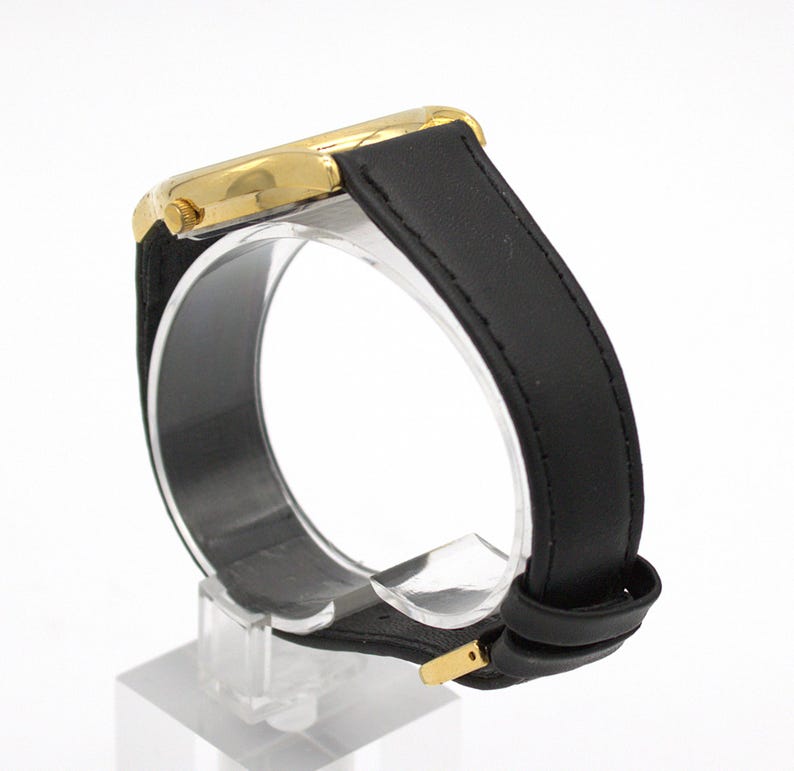 May include: A gold-toned wristwatch with a black leather band. The watch face is gold, and the band is secured with a gold-colored buckle. The watch is displayed on a clear stand against a white background.