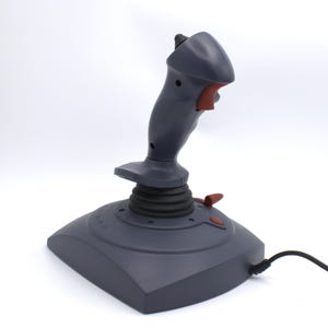Puede incluir: Un joystick gris con detalles en rojo, dise&ntilde;ado para juegos. El joystick tiene una base, una palanca de control central y varios botones. Un cable negro se extiende desde la base. El joystick est&aacute; sobre un fondo blanco.