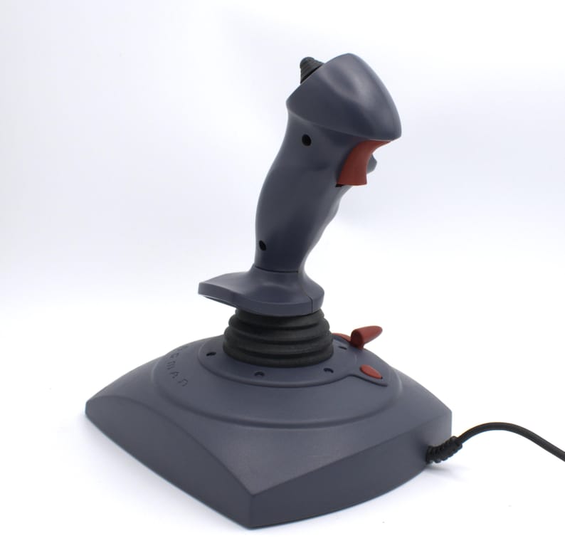 Puede incluir: Un joystick gris con detalles en rojo, dise&ntilde;ado para juegos. El joystick tiene una base, una palanca de control central y varios botones. Un cable negro se extiende desde la base. El joystick est&aacute; sobre un fondo blanco.