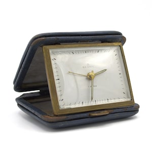 May include: A vintage travel clock with a square face and gold-tone frame, set within a dark blue leather case. The clock face is white with black markings and gold hands. The text "RENSIE" and "7 JEWELS" are visible.