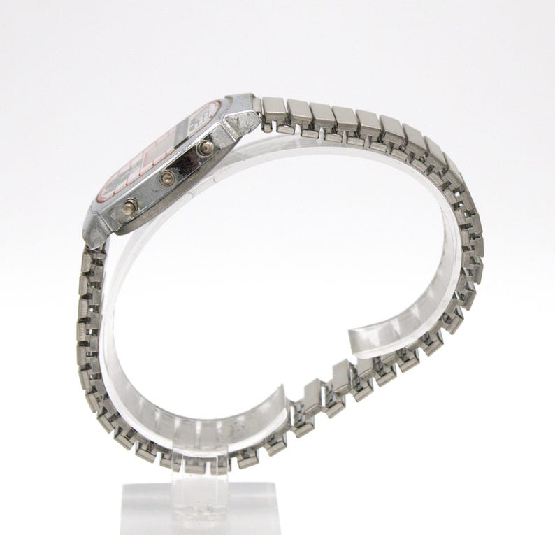 Peut inclure: Une montre-bracelet num&eacute;rique argent&eacute;e avec un cadran rectangulaire et un bracelet en m&eacute;tal &agrave; maillons. Le cadran de la montre a un affichage rouge. La montre est expos&eacute;e sur un support transparent sur fond blanc. La montre est de style vintage.