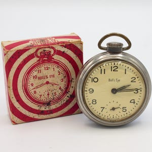 May include: A silver pocket watch with a white face and black hands. The watch face says "Bull's Eye" and has black numbers. The watch is in its original box, which is red and white with a bullseye pattern.