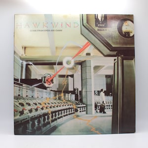 May include: A vintage vinyl record album cover featuring the band Hawkwind. The cover art depicts a futuristic, industrial setting with a man in a white lab coat standing in the foreground. The album title "Quark Strangeness and Charm" is printed in white letters on a black background.