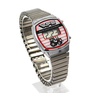 Peut inclure: Une montre num&eacute;rique vintage avec un bracelet argent&eacute; et un cadran rectangulaire. Le cadran affiche le texte "WFE WHITE FARM EQUIPMENT" et un dessin de tracteur. L'&eacute;cran num&eacute;rique affiche l'heure. La montre est sur fond blanc.