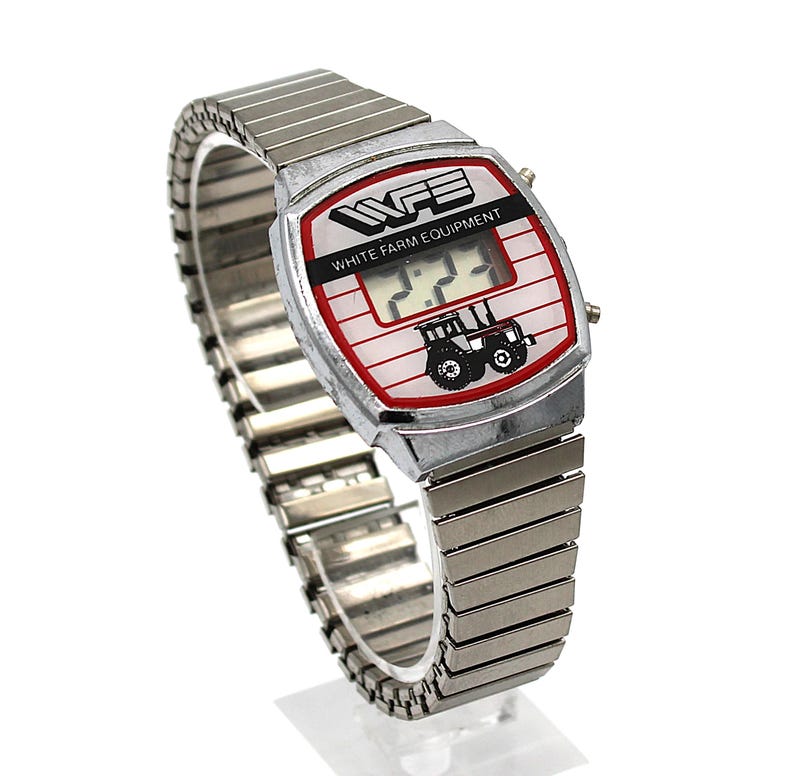 Peut inclure: Une montre num&eacute;rique vintage avec un bracelet argent&eacute; et un cadran rectangulaire. Le cadran affiche le texte "WFE WHITE FARM EQUIPMENT" et un dessin de tracteur. L'&eacute;cran num&eacute;rique affiche l'heure. La montre est sur fond blanc.