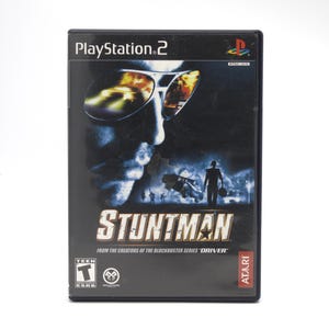 May include: A PlayStation 2 video game case for "Stuntman." The cover features a man's face with sunglasses reflecting a car chase scene. The game title is in large, bold letters, with the Atari logo at the bottom.