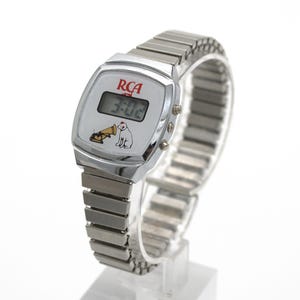 May include: A silver-tone digital wristwatch with a flexible metal expansion band. The watch face features the red RCA logo, a digital time display showing 3:02, and a graphic illustration of a dog sitting next to a gramophone horn.