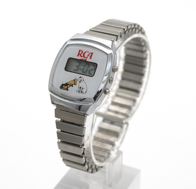 May include: A silver-tone digital wristwatch with a flexible metal expansion band. The watch face features the red RCA logo, a digital time display showing 3:02, and a graphic illustration of a dog sitting next to a gramophone horn.