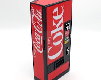 May include: A red and black Coca-Cola vending machine with a silver antenna on top. The machine has a large "Coke" logo on the front and a smaller "Coca-Cola" logo on the side. The machine has a selection of buttons and a coin slot.