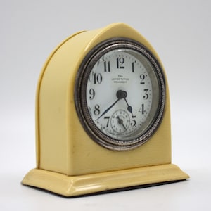 May include: A cream-colored alarm clock with a silver face and black hands. The clock face reads "The Junior Tattoo Movement" and has black Roman numeral hour markers.