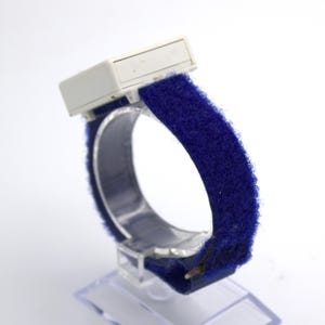 May include: A vintage blue wristband with a white rectangular component. The band is made of a soft, fuzzy material. The watch is displayed on a clear plastic stand, showcasing its design and texture.