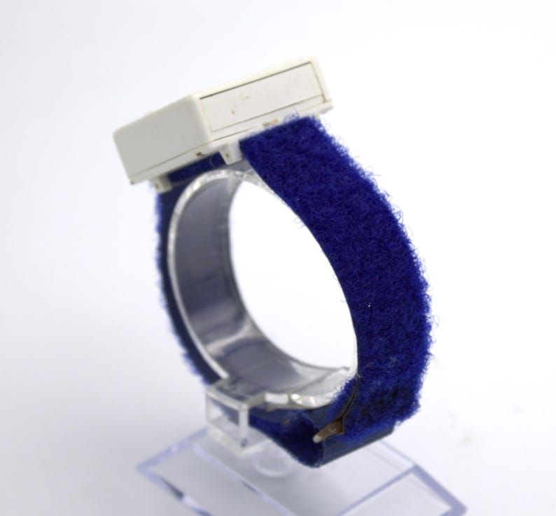 May include: A vintage blue wristband with a white rectangular component. The band is made of a soft, fuzzy material. The watch is displayed on a clear plastic stand, showcasing its design and texture.