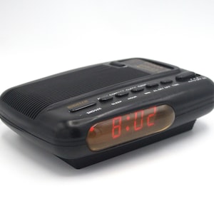 May include: Black digital alarm clock with a red LED display showing the time. The clock has buttons for snooze, sleep, and setting the time. The clock is rectangular and has a ribbed top.
