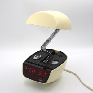 May include: A vintage cream-colored alarm clock with a red digital display showing the time. The clock has a curved top and a chrome adjustable arm. The brand name "Infinity" is printed on the front.