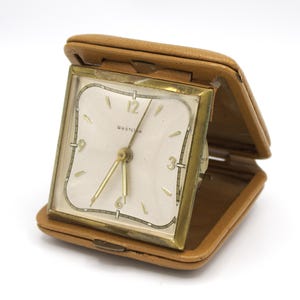 May include: A vintage Westclox travel alarm clock in a tan leather case. The clock face is square with a cream-colored background, gold-tone hands, and a gold-tone frame. The case is open, revealing the clock face.